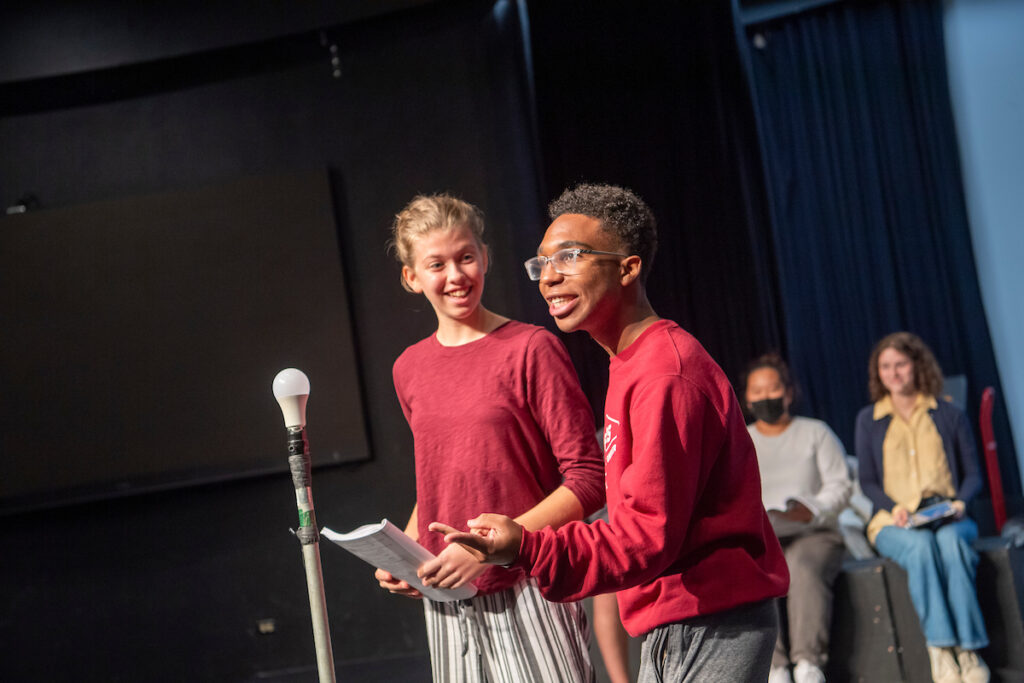 Two students participate in theatre class assignment with skit on stage with microphone